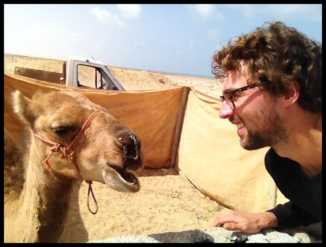 Selfy of Dan looking and grinning at a camel in a small enclosure. The camel has it