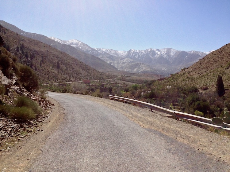 A photo of a road winding up into some mountains. The tallest mountains have snow caps, in the far distance.