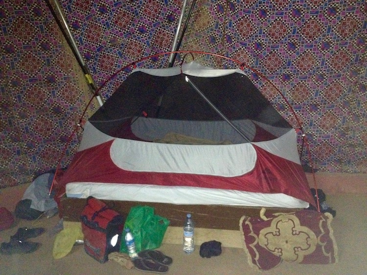 A single bed in a large dorm room, with the inner part of Dan's tent set up on it, and all Dan's belongings sprawled around on the floor.