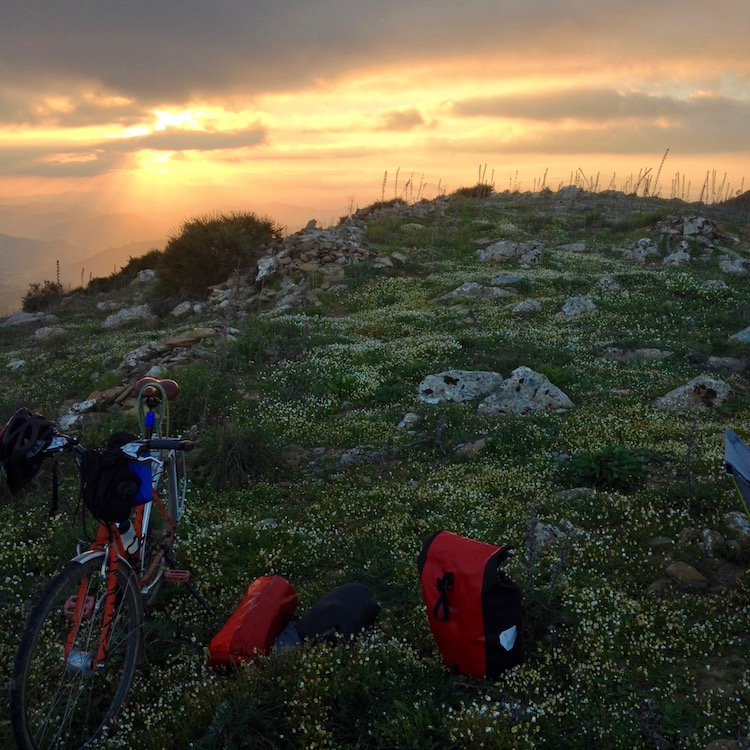 Dan's bike and bags sitting on a patch of soft grass (with daisies) that vanishes into the clouds after a few meters, with the sun breaking through the clouds.