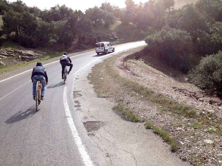 A slightly tilted action shot of two Moroccan kids cycling fast down a hill, turning a bend, with a van also driving in front of them.