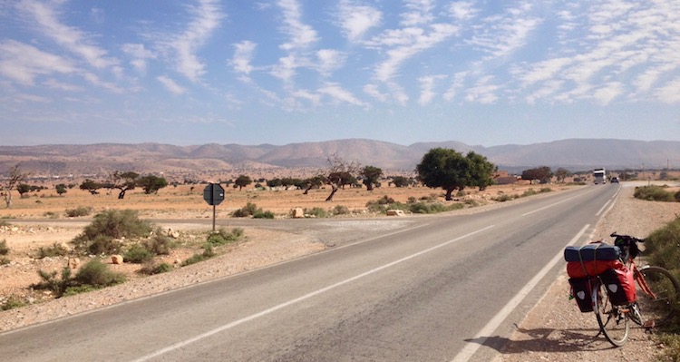 A very straight road stretching across the image and into the distance, with Dan's bike parked beside it. The landscape is mostly sand, but there are small trees scattered around.
