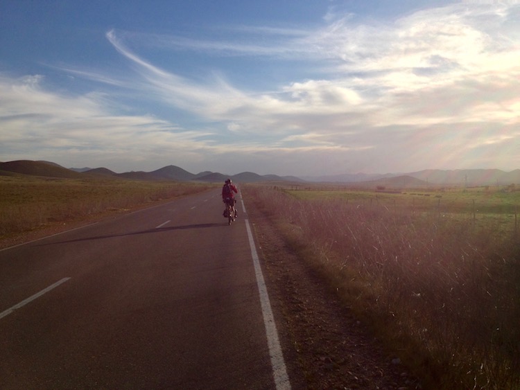 Nico cycling along a straight road surrounded by fields of long grass, and then rolling hills lining the horizon. The setting sun casts long shadows from the grass, sideways across the road.