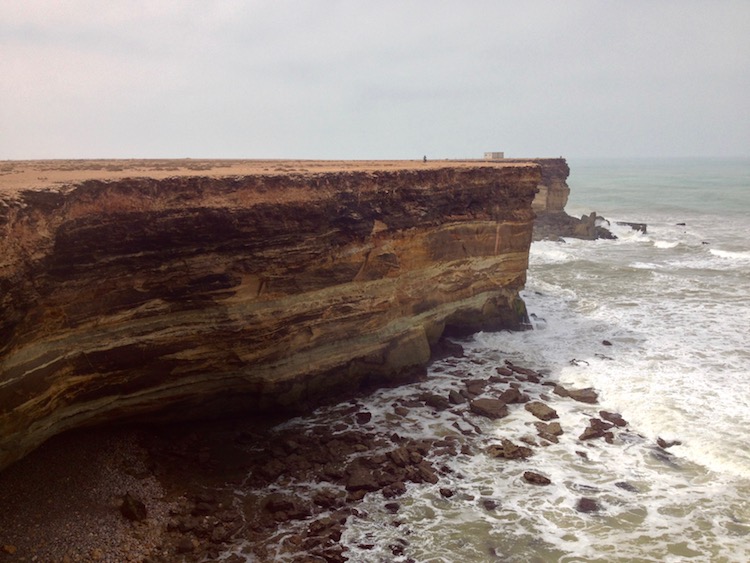 Golden brown cliffs stretching out into the ocean, which is a mix of pale blues, as the waves are crashing against the rocks, blown by the wind. The colour of the sea almost merges into the colour of the sky, which is overcast and grey-blue in colour.