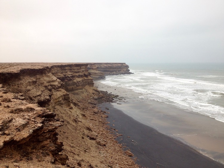 Brown cliffs stretching into the distance and out into the pale blue sea. A beach can be seen below, which, strangely, is coloured black, as if covered in oil.