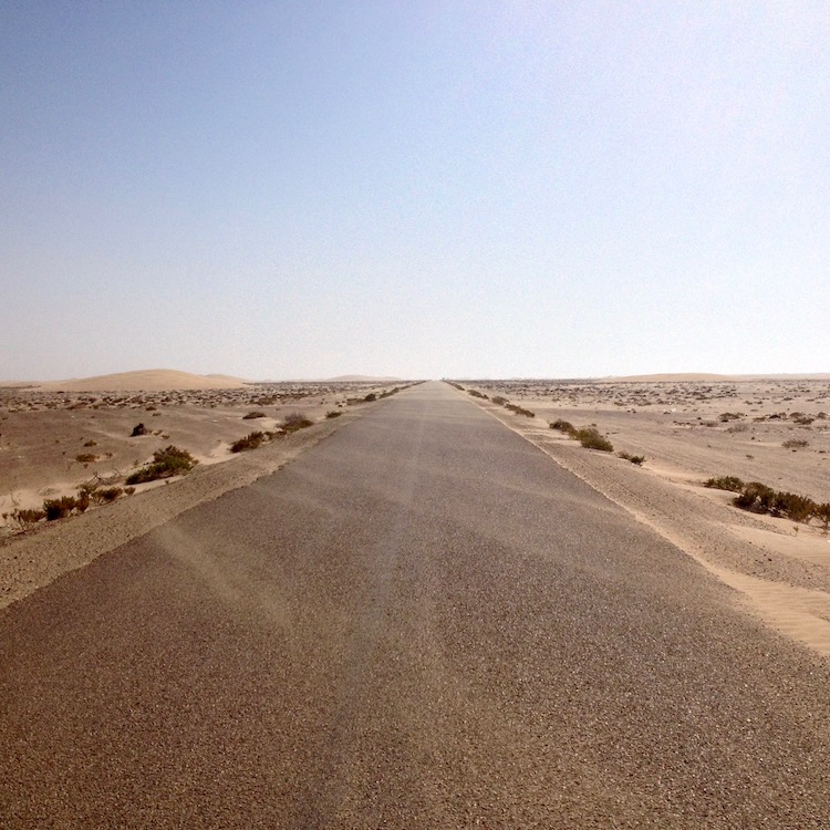 A long, straight, empty road stretching all the way to the horizon, with sand streams blowing diagonally across it. On either side is a desert plain, with the exception of one small sand dune in the distance, on the left side.